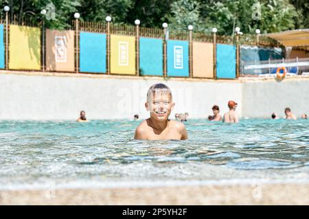 Der fröhliche Schuljunge schwimmt im sauberen blauen Wasser des Pools, schaut in die Kamera und lächelt. Der blonde Junge genießt Spaß im Wasserpark des Sommerresorts Stockfoto