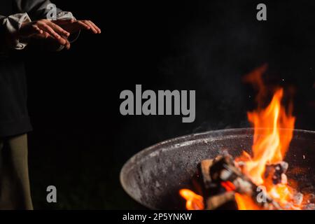 Die Hände des Kindes wärmen sich in der Nähe des Fasses im Dunkeln. Obdachlose, Emigranten, Bettler-Konzept. Stockfoto