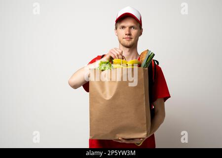 Happy Delivery Mann in roter Uniform mit Kappe und thermische Tasche Rucksack hält braune Bastelpapiertasche mit Lebensmitteln isoliert auf grauem Hintergrund Studio Stockfoto