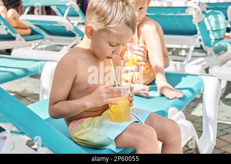 Blonde Brüder genießen erfrischende Getränke mit Strohhalmen, die auf einem Liegestuhl sitzen. Jungs entspannen sich nach Aktivitäten im Wasserpark bei der Schließung des Sommerresorts Stockfoto