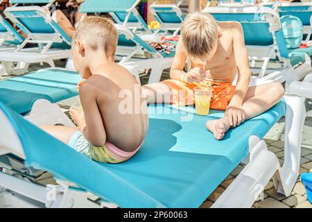 Blonde Brüder genießen erfrischende Getränke mit Strohhalmen, die auf einem Liegestuhl sitzen. Jungs entspannen sich nach Aktivitäten im Wasserpark bei der Schließung des Sommerresorts Stockfoto