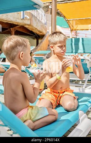 Blonde Brüder genießen erfrischende Getränke mit Strohhalmen, die auf einem Liegestuhl sitzen. Jungs entspannen sich nach Aktivitäten im Wasserpark bei der Schließung des Sommerresorts Stockfoto