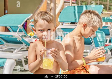 Blonde Brüder genießen erfrischende Getränke mit Strohhalmen, die auf einem Liegestuhl sitzen. Jungs entspannen sich nach Aktivitäten im Wasserpark bei der Schließung des Sommerresorts Stockfoto