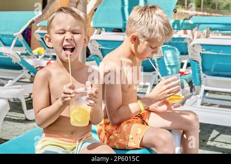 Blonde Brüder genießen erfrischende Getränke mit Strohhalmen, die auf einem Liegestuhl sitzen. Jungs entspannen sich nach Aktivitäten im Wasserpark bei der Schließung des Sommerresorts Stockfoto