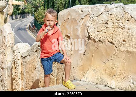 Süßer kleiner Junge knabbert an sonnigen Tagen in der Nähe eines rauen Kalksteinzauns an Plastikhalmen. Ihr Kind posiert während des Urlaubs vor der Kamera Stockfoto