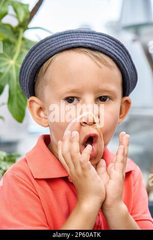 Kleinkinder mit panama-Grimaces und Gummiarmband im Gesicht in der Nähe exotischer Pflanzen. Das verspielte kleine Kind hat Spaß daran, in die Kamera zu schauen Stockfoto