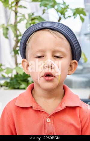 Kleinkinder mit panama-Grimaces und Gummiarmband im Gesicht in der Nähe exotischer Pflanzen. Das verspielte kleine Kind hat Spaß daran, in die Kamera zu schauen Stockfoto