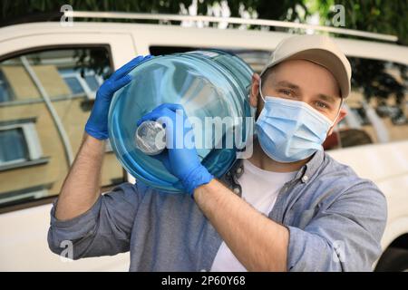 Kurier in medizinischer Maske hält Flasche Kühlwasser in der Nähe des Autos im Freien. Entbindung während der Coronavirus-Quarantäne Stockfoto