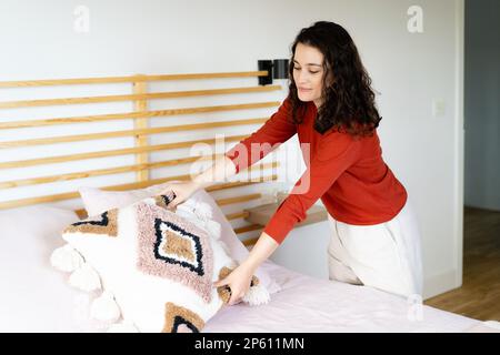 Junge glückliche Frau in rotem Pullover, die zu Hause ein Bett mit Bo-Ho-Kissen macht und dekoriert Stockfoto