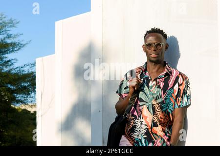 Ein junger gutaussehender Mann afrikanischer Herkunft, der eine Sonnenbrille, Sommerkleidung und einen Rucksack auf dem Rücken trägt, steht gegen ein weißes Panel Stockfoto
