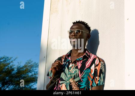 Ein nachdenklicher junger Mann afrikanischer ethnischer Herkunft, trägt eine Sonnenbrille, kurzes Haar, ruht sich aus, analysiert und blickt in den Horizont. Stockfoto
