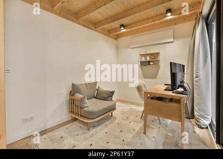 Ein Wohnzimmer mit weißen Wänden und Holzdecken, es gibt eine graue Couch vor dem Fenster Stockfoto