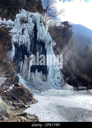 Eine atemberaubende Landschaft mit einem Fluss, der sich durch ein felsiges Gelände schlängelt, hervorgehoben durch einen herrlichen gefrorenen Wasserfall, der den felsigen Hang hinunterstürzt Stockfoto