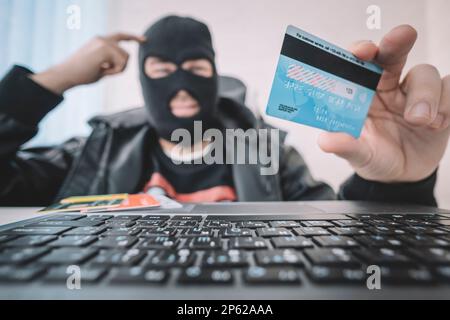 Internetdiebstahl - ein Mann, der eine Balaklava trägt und eine Kreditkarte hält, während er hinter einem Laptop sitzt, ist ein Konzept, Geld über das Internet zu stehlen. Kredit c Stockfoto