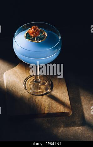 Ein schönes, abgerundetes Glas mit blauer Flüssigkeit, ein trockenes Stück Zitrone, eine trockene Blume und eine dunkle Noir-Stimmung mit starkem Schatten. Stockfoto