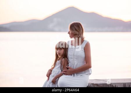 Schwangere Mutter der Millennials sitzt mit ihrer Tochter bei Sonnenuntergang am Meer. Eine Frau mittleren Alters umarmt ein kleines Mädchen. Die Reise alleinerziehender Eltern. Gesunde Schwangerschaft Stockfoto