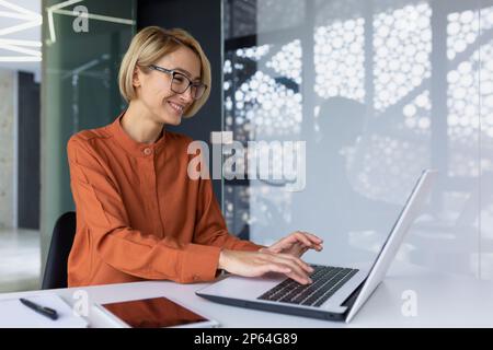 Wunderschöne junge Geschäftsfrau im Büro, die mit einem Laptop arbeitet, blond mit kurzen Haaren, die auf der Tastatur tippen und lächeln, weibliche Programmiererin, die Software programmiert, während sie am Arbeitsplatz sitzt. Stockfoto