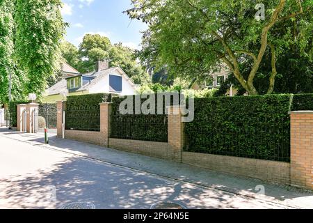 Amsterdam, Niederlande - 10. April 2021: Ein Backsteinzaun vor einem Haus mit Bäumen auf beiden Seiten und Büschen, die am Straßenrand wachsen Stockfoto