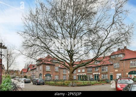 Ein großer Baum in der Mitte einer Straße mit Autos, die auf beiden Seiten geparkt sind, und Gebäuden dahinter, unter einem hellblauen Himmel Stockfoto