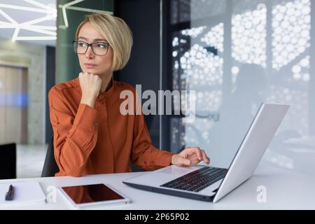 Eine Geschäftsfrau, die im Büro mit einem Laptop aus dem Fenster schaut, trifft eine blonde Angestellte mit Brille eine Entscheidung über die Finanzen des Unternehmens. Stockfoto