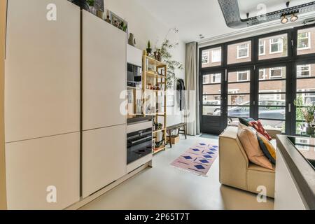 Amsterdam, Niederlande - 10. April 2021: Küche und Wohnzimmer in einem Apartment mit viel Tageslicht, das durch die großen Glastüren an den Fenstern strömt Stockfoto