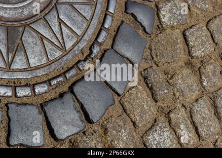 Alte Abwasserluke aus Metall in der Mitte der Straße, aus einem glatten Pflasterstein. Stockfoto