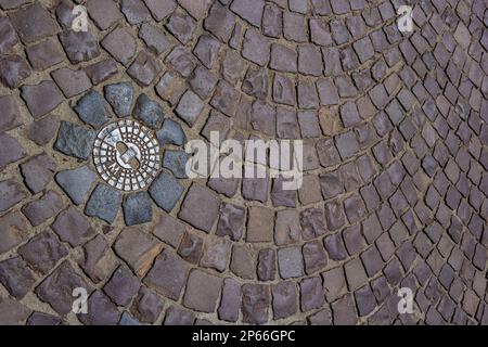 Alte Abwasserluke aus Metall in der Mitte der Straße, aus einem glatten Pflasterstein. Stockfoto