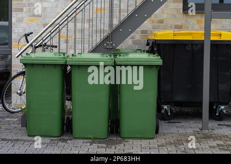 Drei grüne Mülltonnen vor einem Wohngebäude. Fahrrad und schwarzer Abfalleimer unter der Metalltreppe. Stockfoto