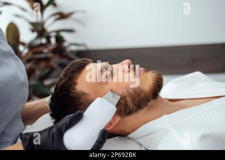 Die Schönheitsmeisterin macht Ultraschall-Gesichtspeeling für ihren Klienten. Youngkaukasischer Mann, der in einem Schönheitssalon Haut und Poren effektiv reinigt. Stockfoto