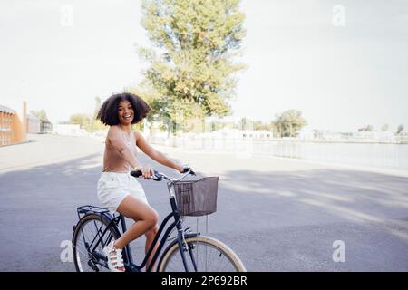 Eine dunkelhäutige Teenagerin fährt ihr Fahrrad. Ein junges und positives afro-amerikanisches Mädchen in lässiger Kleidung lächelt süß und hat Spaß. Stadtstraße an Stockfoto