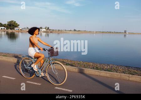 Eine dunkelhäutige Teenagerin fährt ihr Fahrrad. Ein junges und positives afro-amerikanisches Mädchen in lässiger Kleidung lächelt süß und hat Spaß. Stadtstraße an Stockfoto