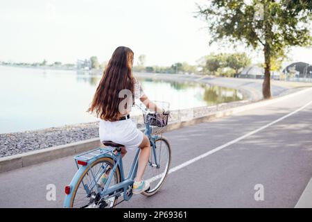 Rückansicht einer Teenagerin fährt ihr Fahrrad. Ein junges und positives Mädchen in lässiger Kleidung hat Spaß. Stadtstraße und Bäume mit Büschen im Hintergrund Stockfoto