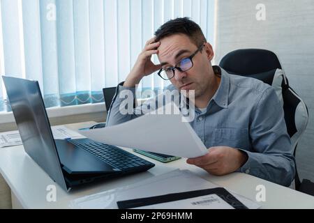 Ein Geschäftsmann, der von der Buchhaltung in seinem Büro deprimiert wird. Ein Mitarbeiter ist überrascht, ein Papierdokument zu lesen, während er am Arbeitsplatz sitzt. Stockfoto