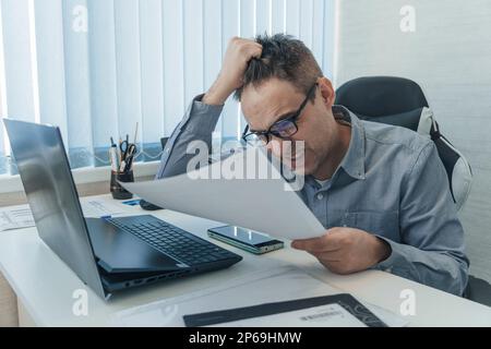 Der junge gestresste Geschäftsmann überwältigte ihn mit Dokumenten auf seinem Schreibtisch, die seinen Kopf nach unten blickten. Müdigkeit und Stau am Arbeitsplatz. Entlassung. Stockfoto