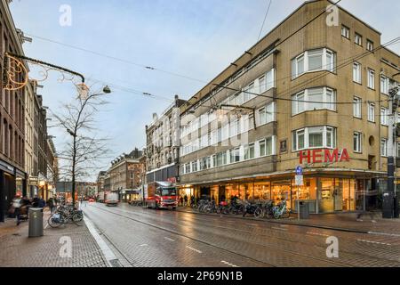 Amsterdam, Niederlande - 10. April 2021: Eine Stadtstraße mit Menschen, die auf Gehwegen unterwegs sind, und Autos, die die Straße vor den Gebäuden in der Abenddämmerung entlang fahren Stockfoto