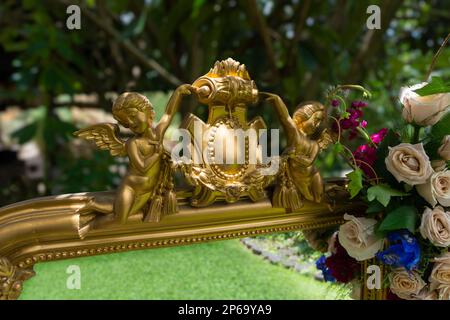 Details der goldenen Engel eines antiken Spiegels, geschmückt mit Rosen in der Mitte des Gartens, reflektiert das Gras mit einem Hintergrund, der unscharf ist Stockfoto