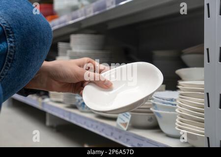 Nahaufnahme einer Frauenhand, die eine weiße Schüssel in einem Baumarkt hält. Verschiedene Platten, die in Regalen in einem Baumarkt ausgestellt werden. Stockfoto