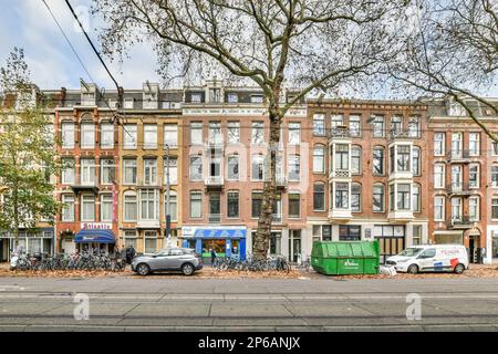Amsterdam, Niederlande - 10. April 2021: Eine Stadtstraße mit Autos, die an der Seite geparkt sind, und Gebäuden im Hintergrund, die alle mit Blättern bedeckt sind Stockfoto