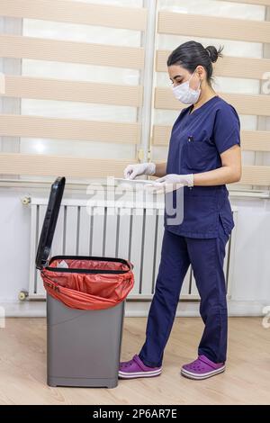 Krankenschwester wirft medizinischen Müll in medizinische Abfalleimer im Büro. Eine Krankenschwester mit einem roten Müllsack. Dienstmädchen und Infektionsmüll im Krankenhaus. Infecti Stockfoto