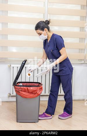 Krankenschwester wirft medizinischen Müll in medizinische Abfalleimer im Büro. Eine Krankenschwester mit einem roten Müllsack. Dienstmädchen und Infektionsmüll im Krankenhaus. Infecti Stockfoto