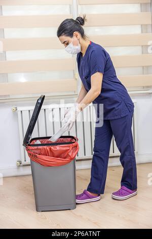 Krankenschwester wirft medizinischen Müll in medizinische Abfalleimer im Büro. Eine Krankenschwester mit einem roten Müllsack. Dienstmädchen und Infektionsmüll im Krankenhaus. Infecti Stockfoto