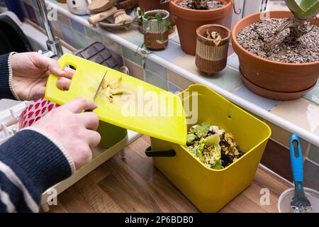Mann, der Lebensmittelabfälle aus einem Schneidebrett in einen Behälter für Lebensmittelrecycling in einer Hausküche schiebt, bereit für die Kompostierung, Großbritannien. Thema: Nachhaltigkeit zu Hause Stockfoto
