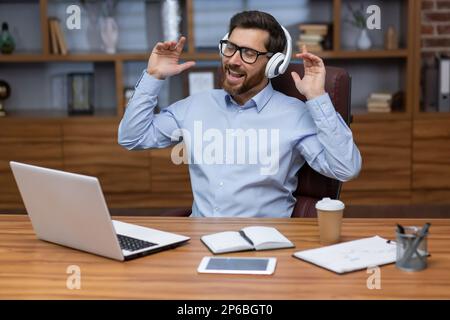 Ein junger Geschäftsmann sitzt im Büro am Tisch, arbeitet an einem Laptop mit weißen Kopfhörern und hört Musik, ruht sich aus. Singt mit, tanzt. Stockfoto
