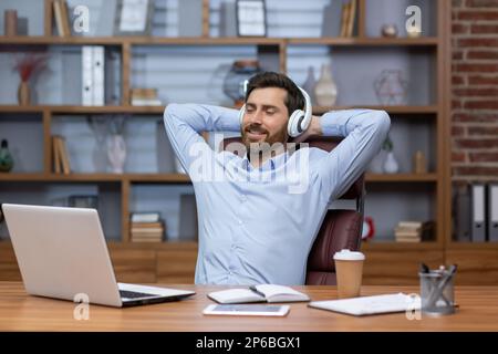 Ein junger männlicher Geschäftsmann sitzt im Büro auf einem Stuhl am Schreibtisch. Er legte die Hände hinter den Kopf, hörte Musik in weißen Kopfhörern, schloss die Augen, ruhte sich aus. Stockfoto
