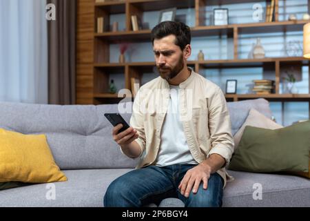 Kommunikationsprobleme. Ein seriöser junger Mann in einem beigen Hemd sitzt zu Hause auf dem Sofa, hält das Telefon, sieht besorgt auf den Bildschirm, schreibt eine Nachricht. Stockfoto