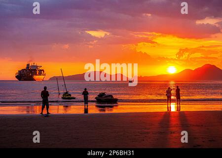 Sonnenuntergang mit rötlichem Himmel an den Stränden von Santos, São Paulo, Brasilien. Frachtschiff und Menschen, die über das Meer nachdenken. Stockfoto