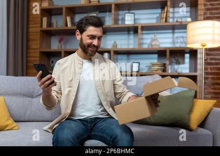 Ein junger Mann sitzt zu Hause auf dem Sofa, hält ein Telefon in der Hand, freut sich, ein Paket bei der Lieferung erhalten zu haben und öffnet die Schachtel. Stockfoto