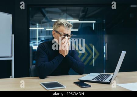 Erwachsener, erwachsener Geschäftsmann am Arbeitsplatz, krank, arbeitet an einem Laptop, grauhaariger Mann niest. Stockfoto
