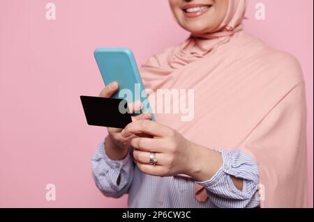 Schwarze Kreditkarte in den Händen einer muslimischen Frau, die ein Smartphone benutzt und die einfache, schnelle und sichere Überweisung von Geld über eine Bank-App genießt. Stockfoto
