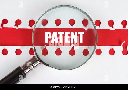 Zwischen zwei Blättern aus einem Notizbuch auf einem roten Streifen mit der Aufschrift „Patent“ befindet sich eine Lupe. Stockfoto